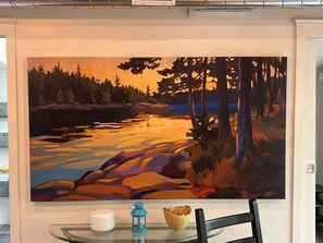 Una pintura que muestra un paisaje forestal con un lago, una silla, una pequeña mesa con una vela y un farol, y una lámpara fijada a la pared.