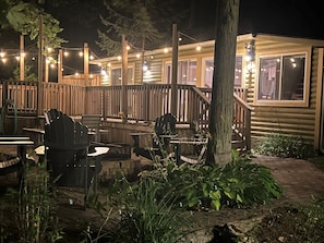 Une terrasse en bois avec des siĂšges dâextĂ©rieur, un foyer en pierre et des guirlandes lumineuses.