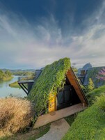 Una casa moderna con techo inclinado cubierto de vegetación, una terraza de madera y vistas a un lago y montañas.