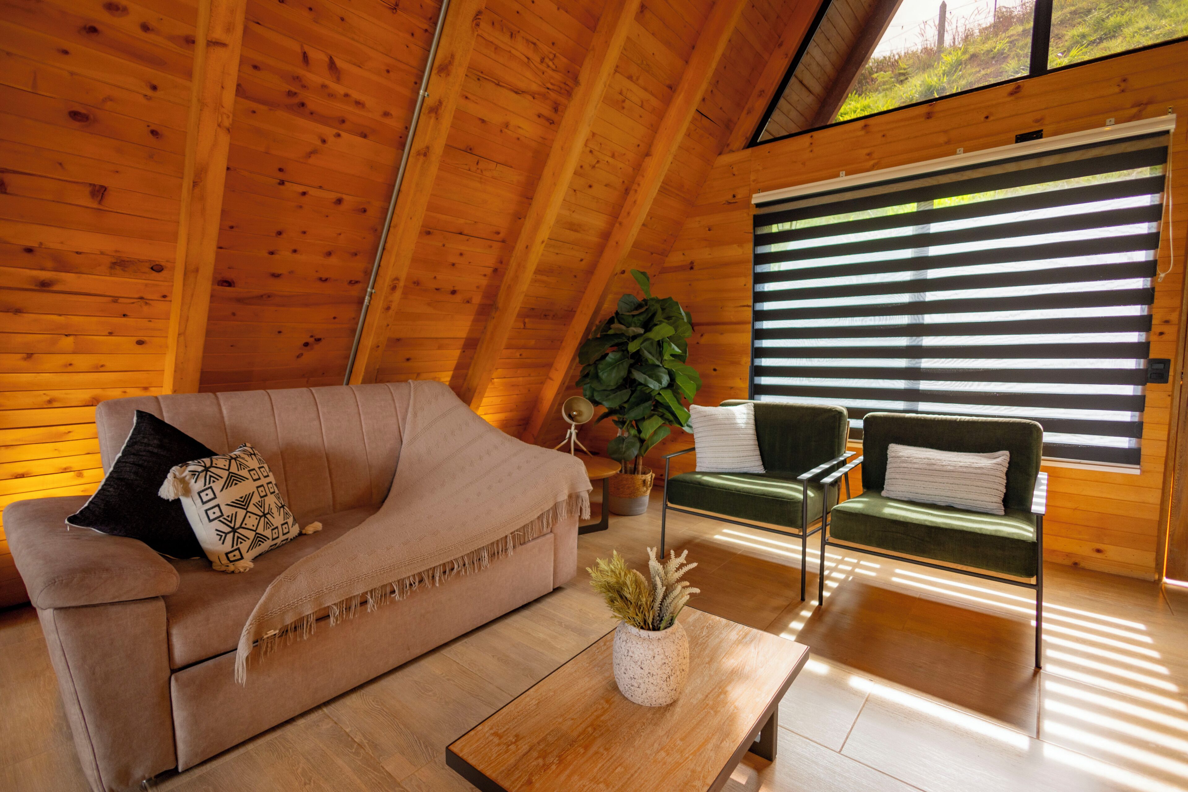 A cozy room with a wooden ceiling, a sofa, a chair, a coffee table, and a large window with blinds.