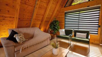 A cozy room with a wooden ceiling, a sofa, a chair, a coffee table, and a large window with blinds.