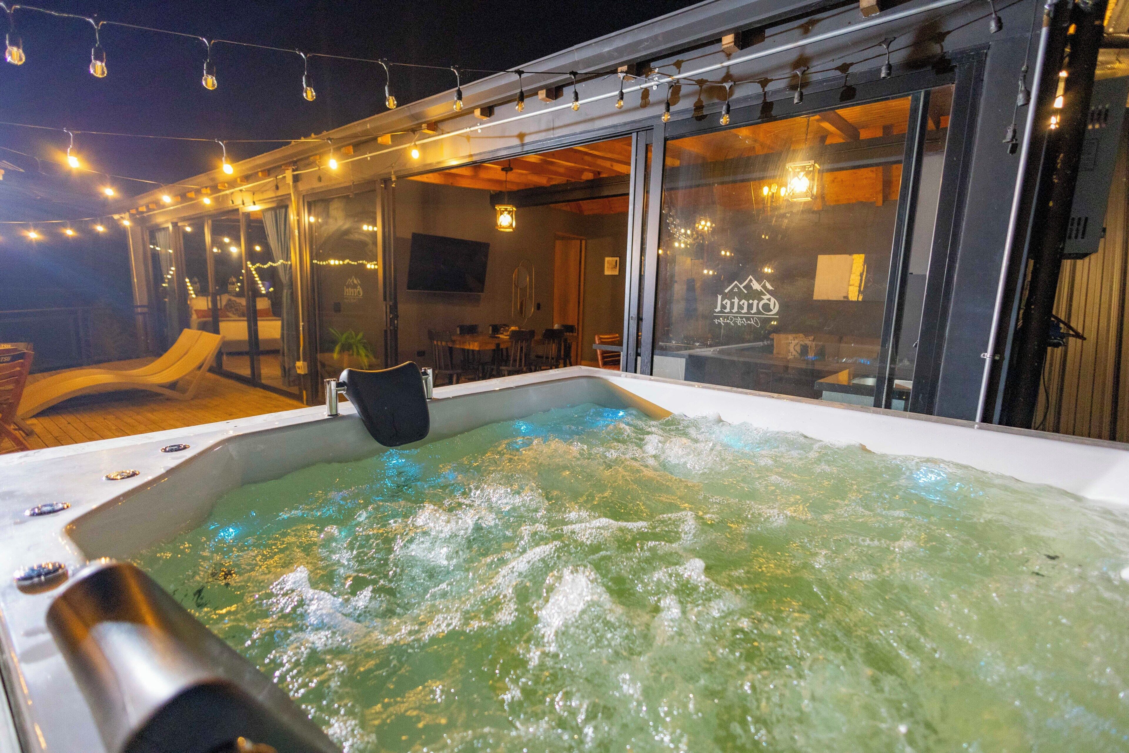 Un jacuzzi con cerramiento de vidrio, rodeado por una terraza con mobiliario exterior e iluminación colgante.