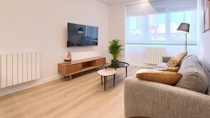 Un salon moderne doté d’un téléviseur à écran plat, d’une console en bois, d’une table basse ronde, d’un canapé gris et d’une grande fenêtre offrant une vue sur les immeubles.