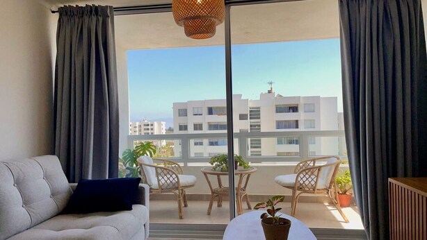 A modern living room with a sofa, a coffee table, and a balcony with wicker chairs and potted plants. - Perfect for digital nomads, families and travelers. Fast WiFi and pools. (La Serena)