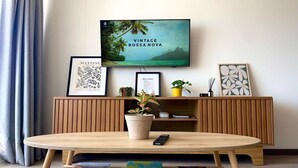 A modern living room with a wooden TV stand, a mounted TV displaying 'Vintage Bossa Nova', a round wooden coffee table with books, a potted plant, and framed artwork. - Perfect for digital nomads, families and travelers. Fast WiFi and pools. (La Serena)