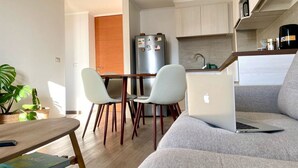 A modern living room with a grey sofa, a wooden dining table with two chairs, a laptop on the sofa, and a kitchen area with a refrigerator and cabinets. - Perfect for digital nomads, families and travelers. Fast WiFi and pools. (La Serena)