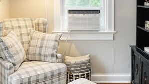 A bedroom with a plaid sofa, a white bed, a small side table, a lamp, and a window with blinds.