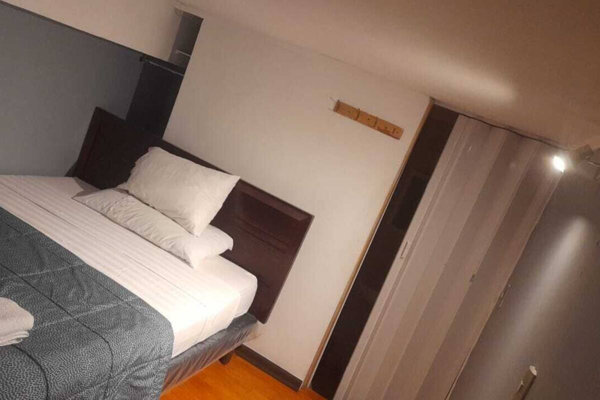 Una cama individual con cabecera oscura, colchón blanco y manta gris.