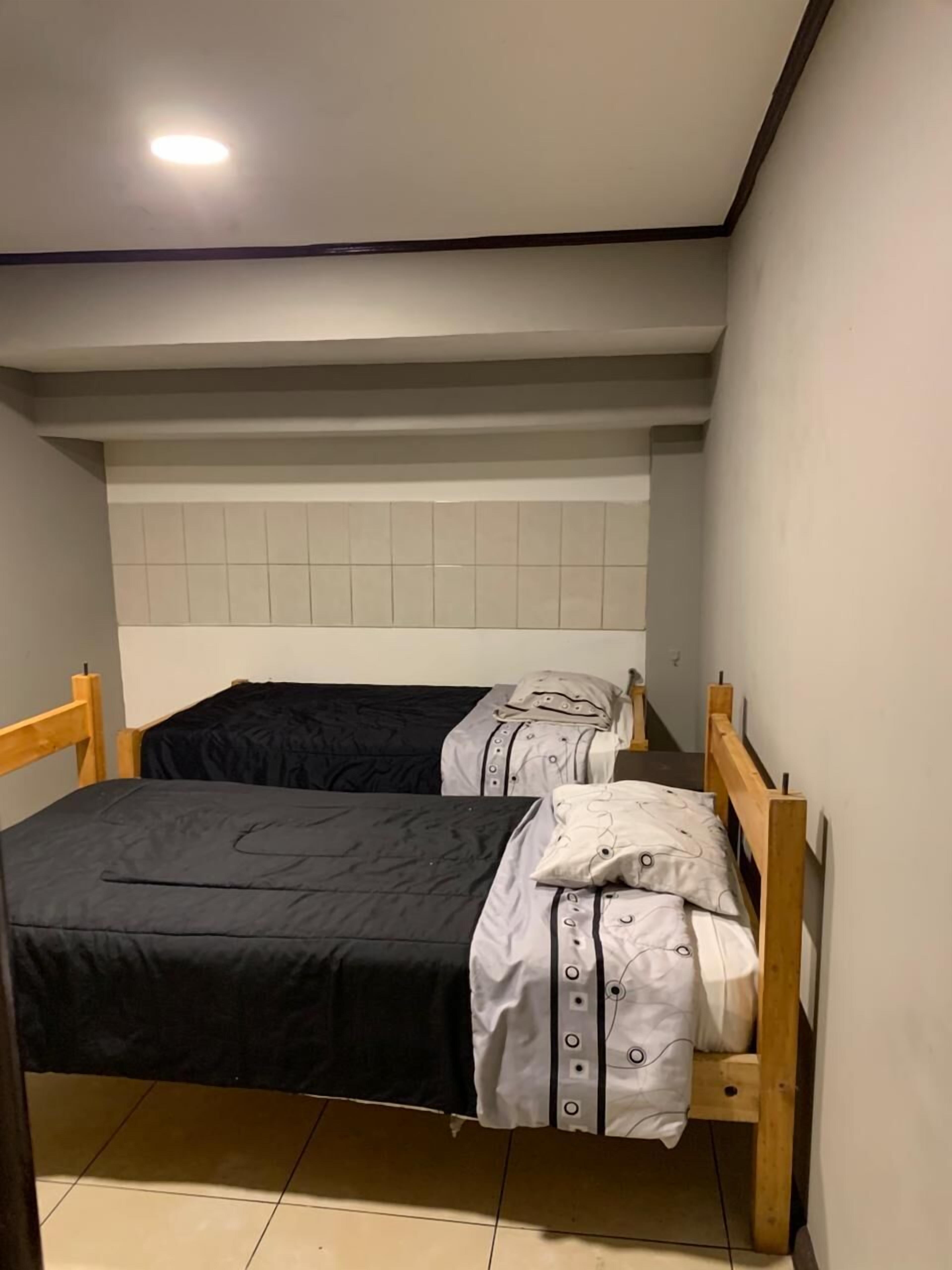 Una habitación pequeña y estrecha con una cama individual, cabecera de madera y pared con azulejos blancos.