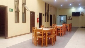 A spacious indoor area with a long row of wooden tables and chairs, a fire extinguisher, a television mounted on the wall, and a sign indicating an exit. - edificio catedral (Santiago)