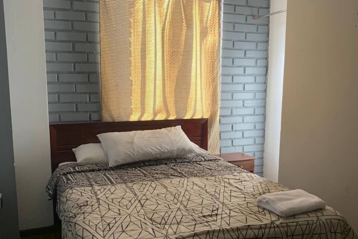 Un dormitorio con cama, cabecera, ventana con cortinas y pared de ladrillo.