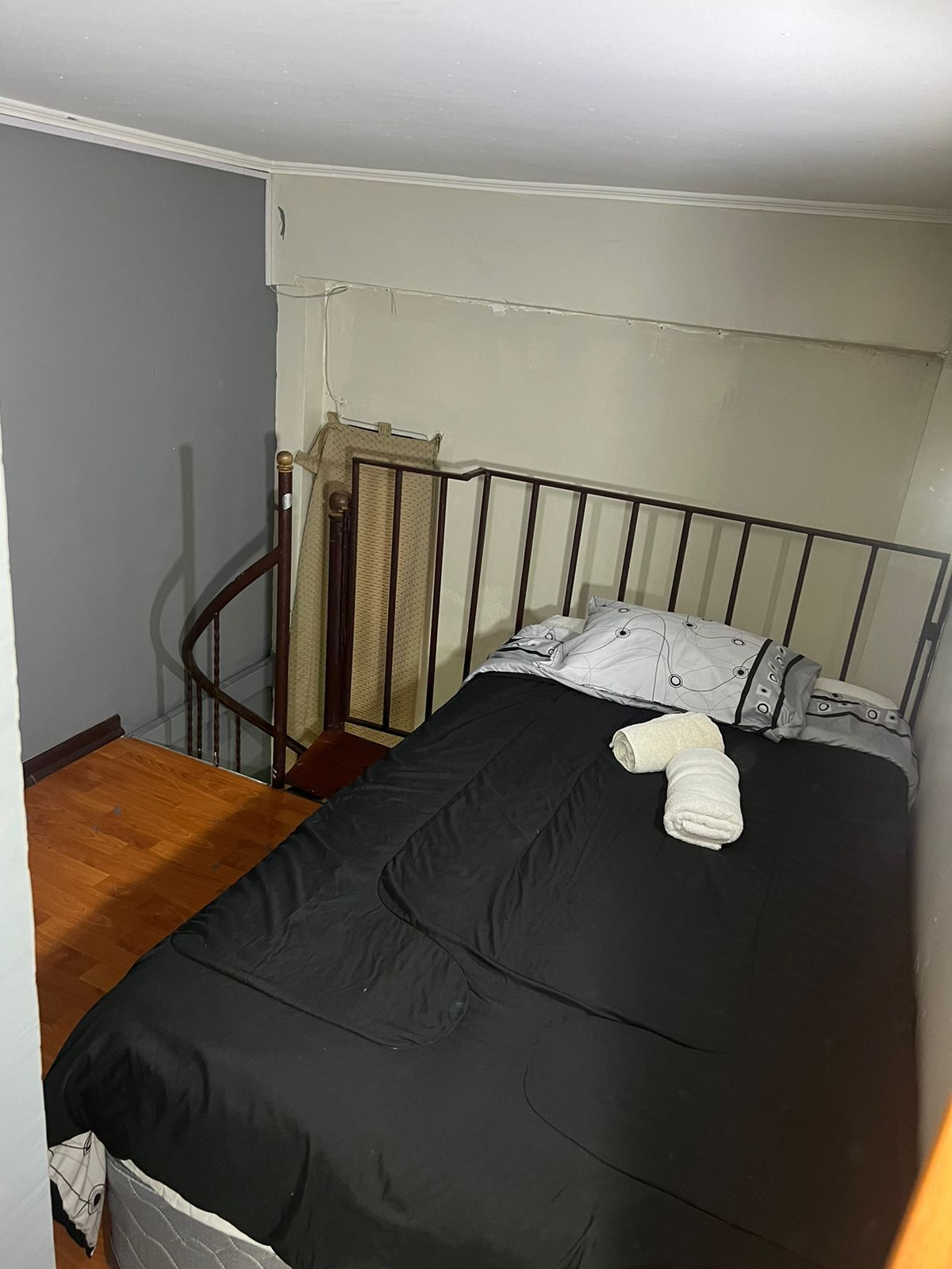 Ein kleines Zimmer mit einem Einzelbett, Holzboden, einem Metall-Kopfteil und einem Fenster mit Vorhang.