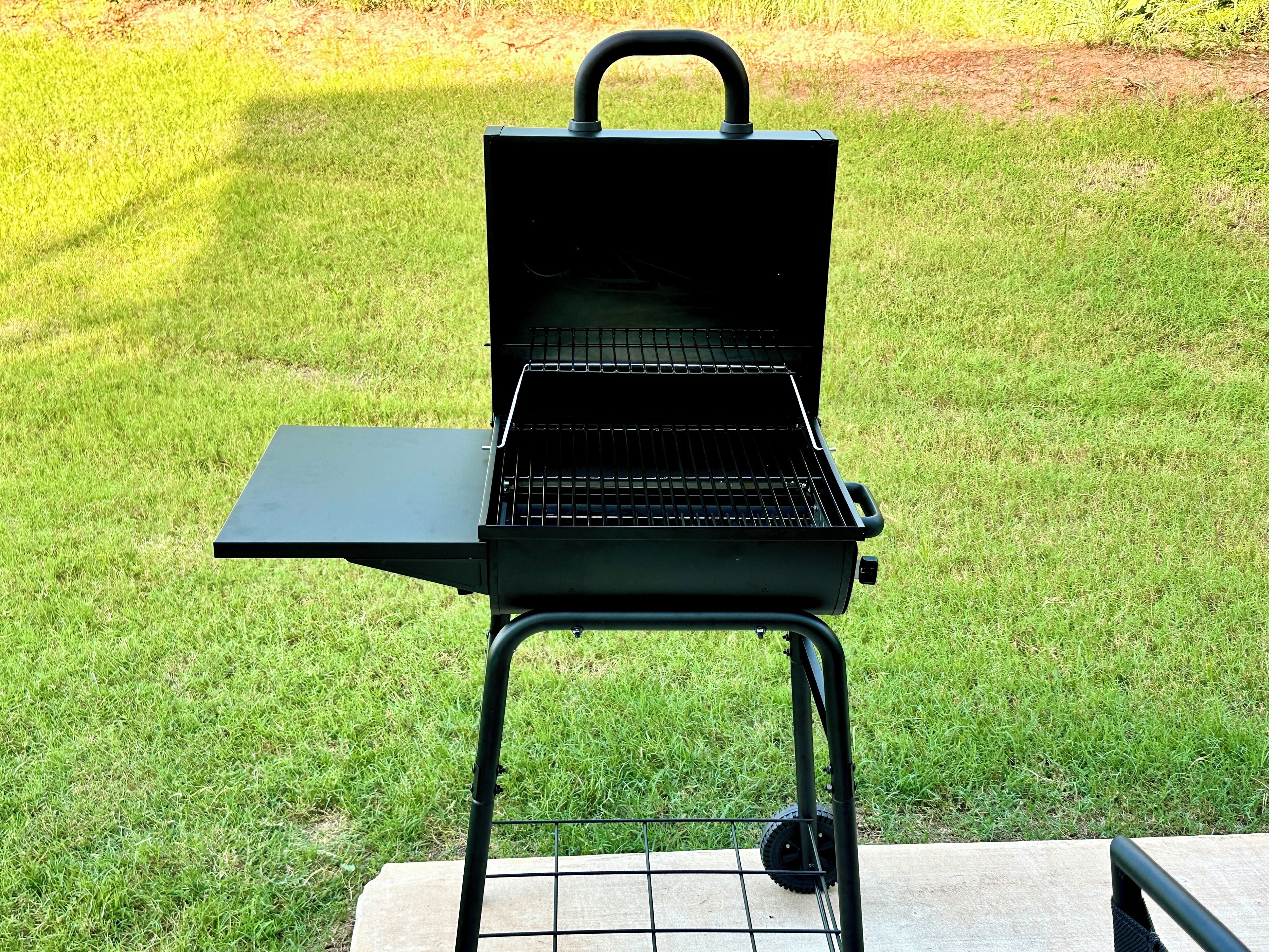 A portable grill with a handle and wheels, placed on a concrete surface in a grassy area.