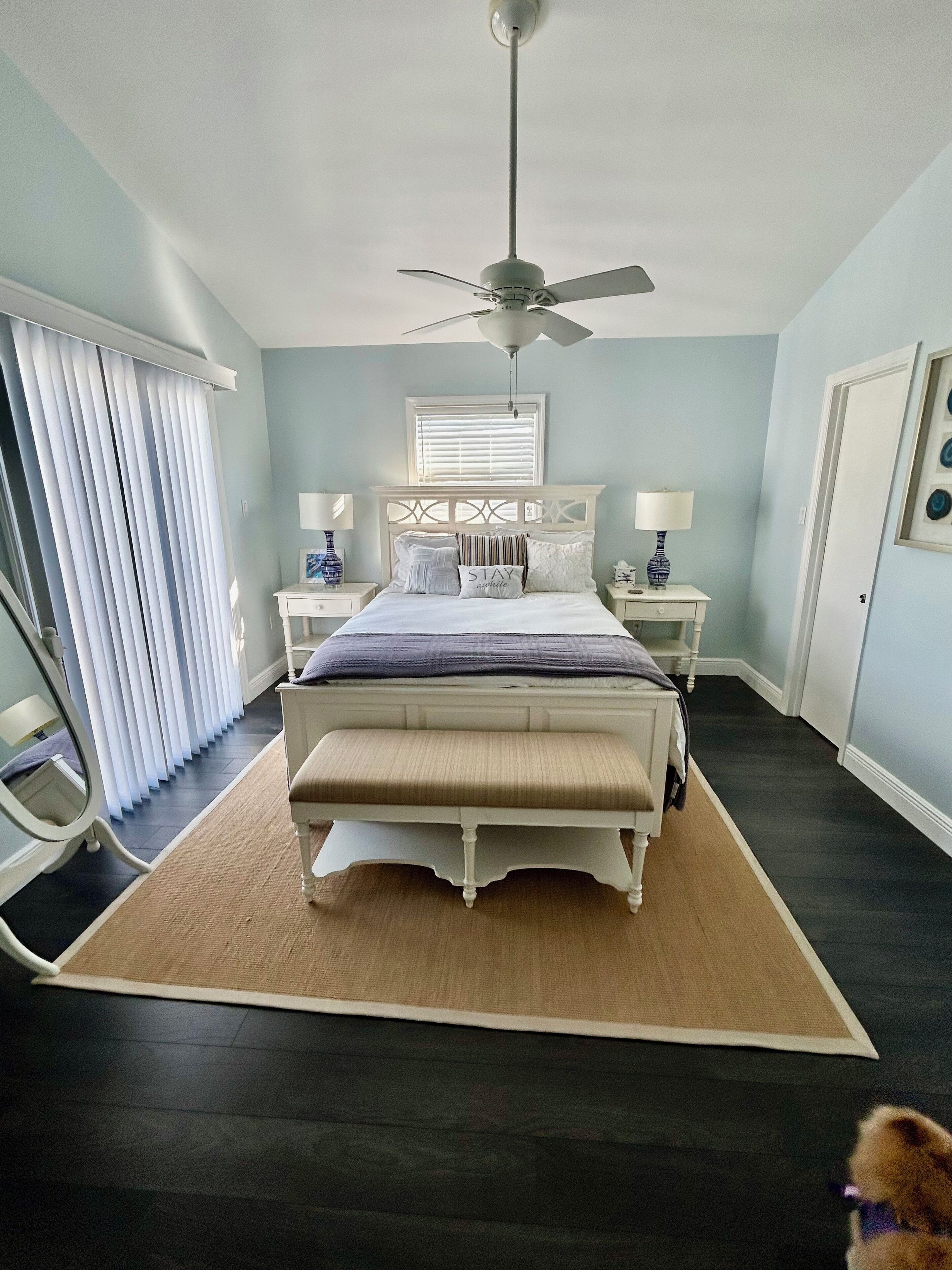 A bedroom with a bed, bedside tables, a ceiling fan, a window with blinds, and a door.