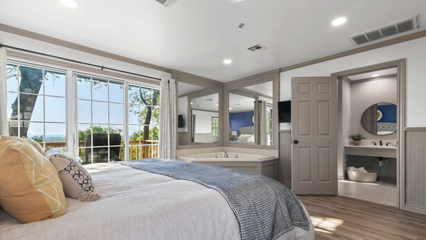 Une chambre avec un grand lit, une fenêtre avec des rideaux, une salle de bain avec un lavabo et un miroir, et une porte donnant sur une autre pièce.