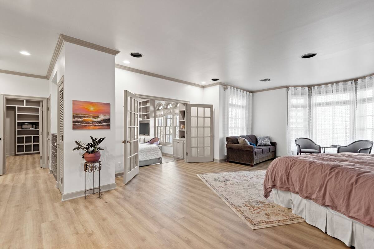 A spacious bedroom with a large bed, a seating area with a sofa and chairs, a walk-in closet, and a view of the outdoors through a window.