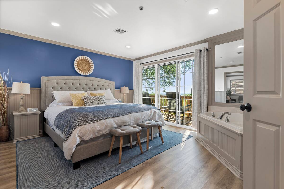 A bedroom with a large bed, a window with a view, a bathroom with a bathtub, and a door to the outside.