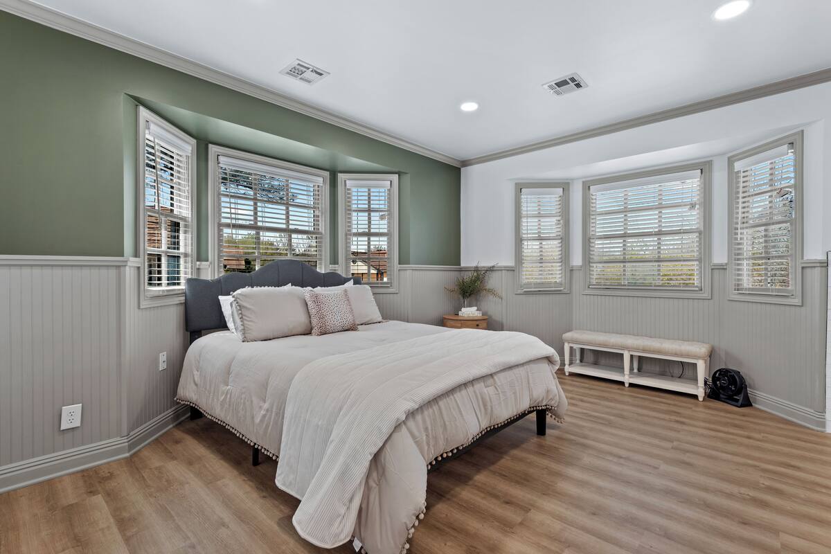 A bedroom with a large bed, a bench, and multiple windows with blinds.