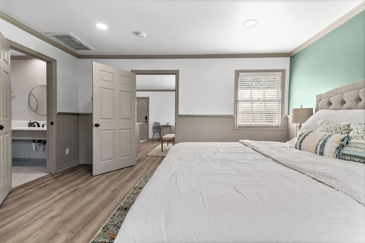A bedroom with a large bed, a dresser, a mirror, and a door leading to another room.