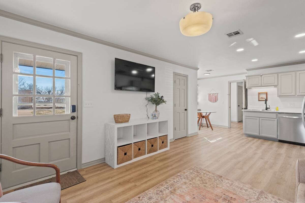 A spacious living area with a wooden floor, a flat-screen TV mounted on the wall, a white console table, and a kitchen area with white cabinets and a countertop.