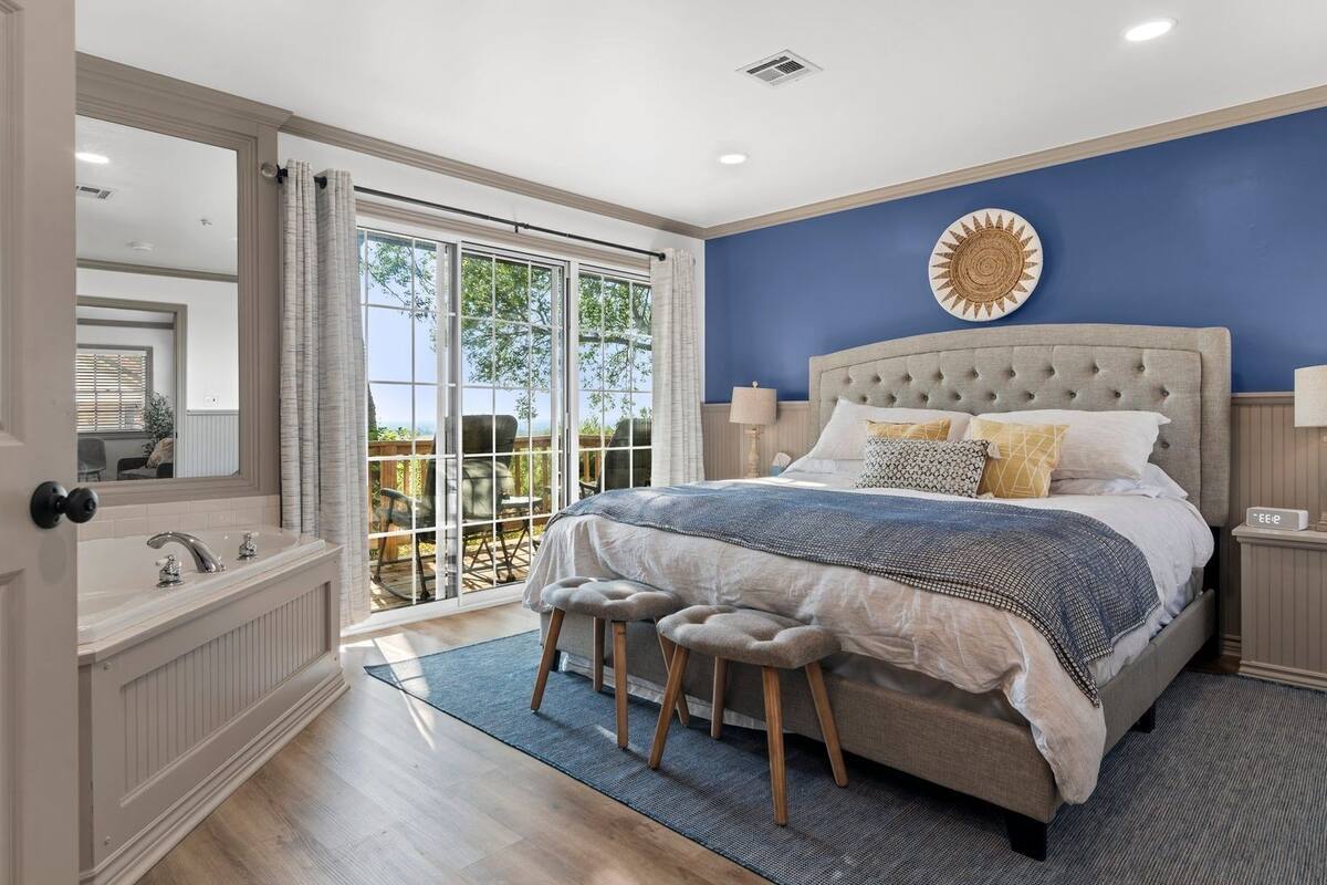 A bedroom with a large bed, a nightstand, a mirror, a window with a view, and a bathroom with a sink and toilet.
