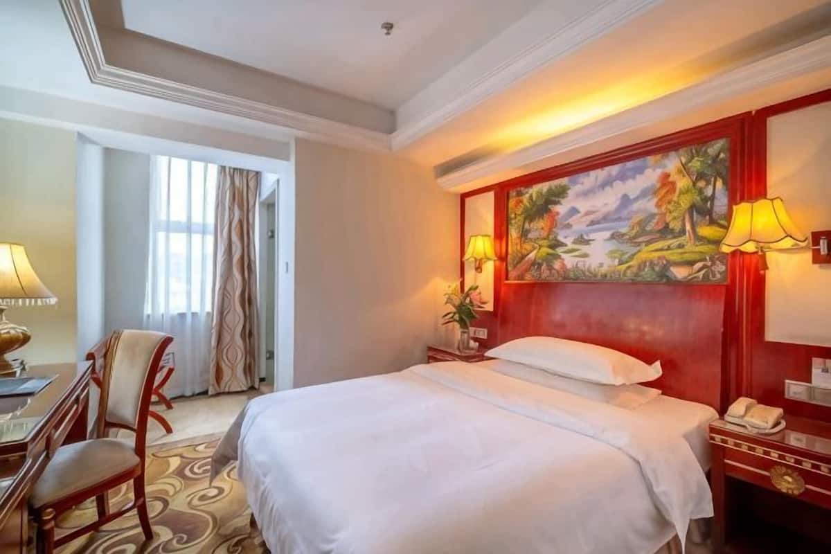 A hotel room with a bed, a desk, a chair, a painting on the wall, and a lamp.