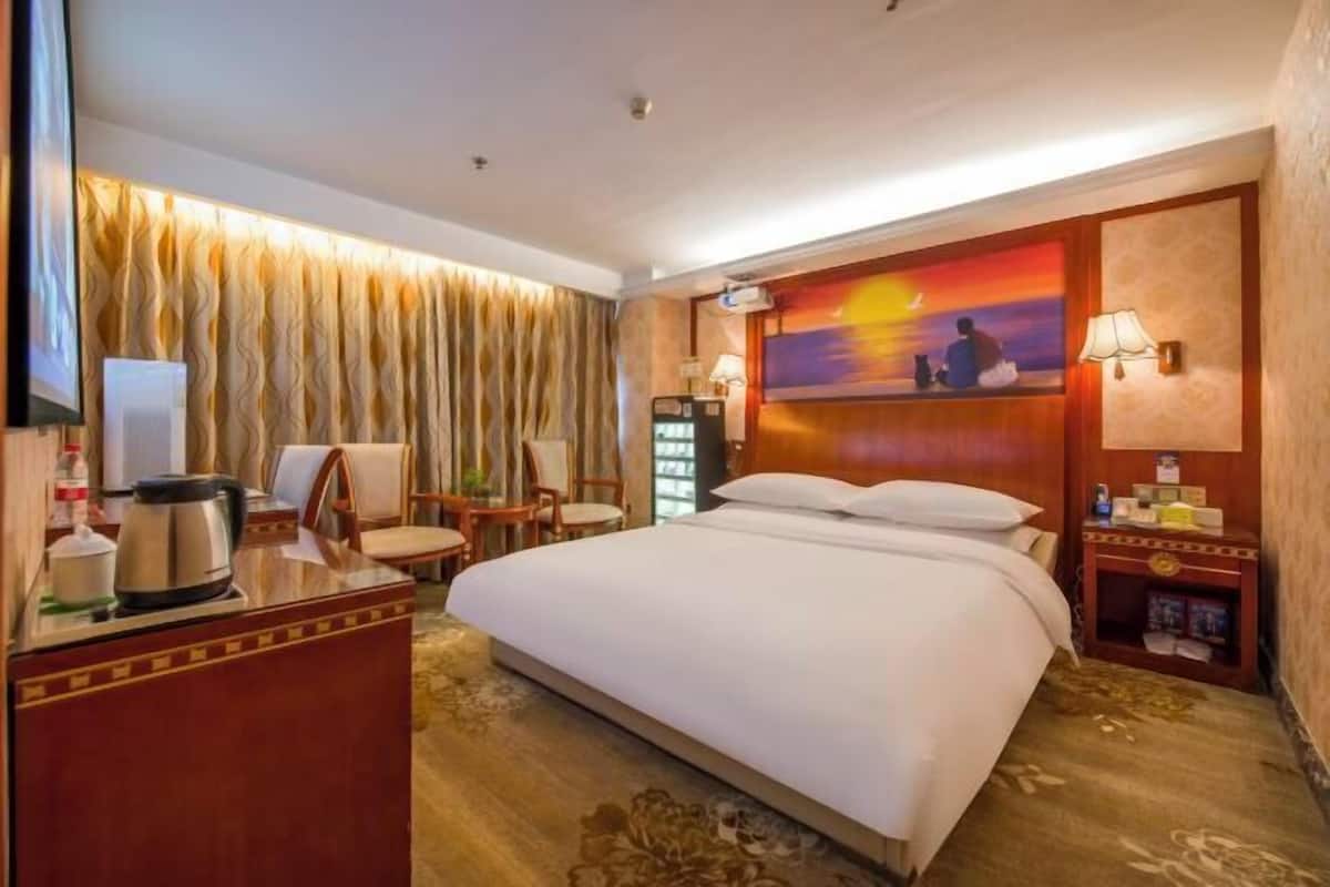 A hotel room with a bed, a desk with a kettle, a chair, a TV, a painting, and a nightstand with a lamp.