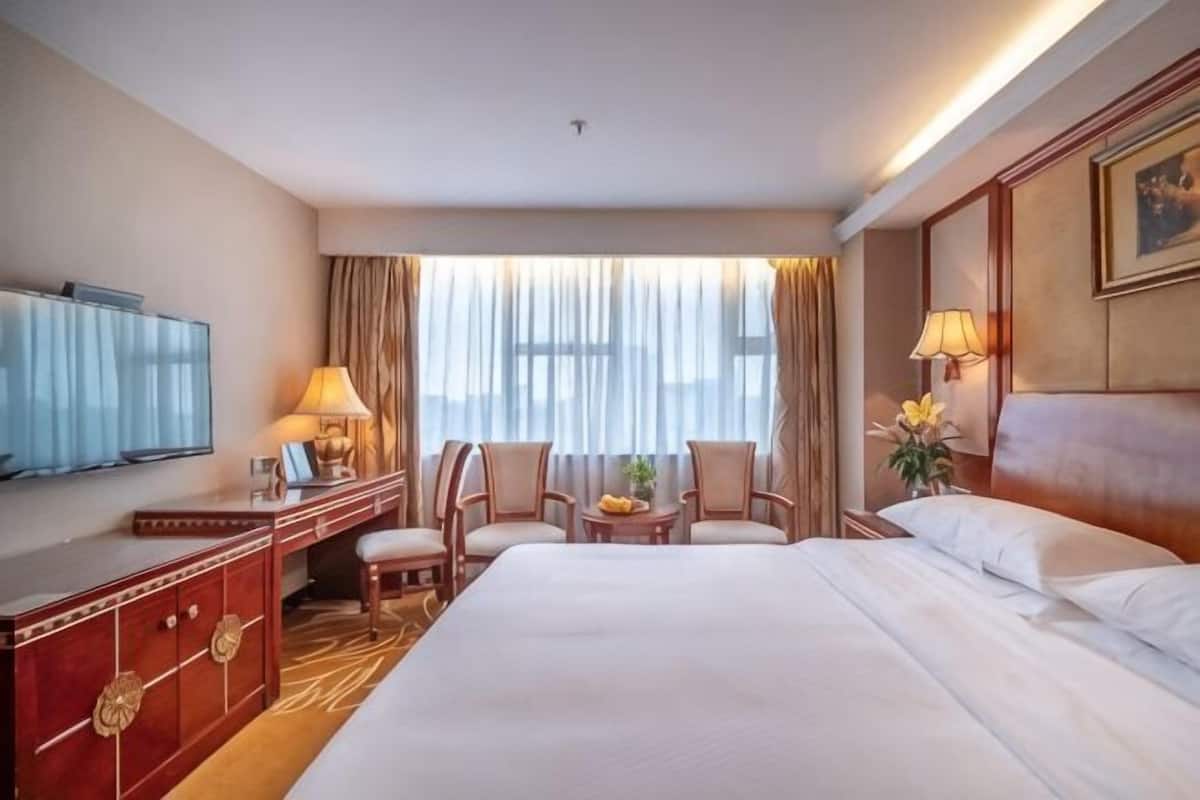 A hotel room with a large bed, a desk with a flat-screen TV, two chairs, a small table, and a window with curtains.