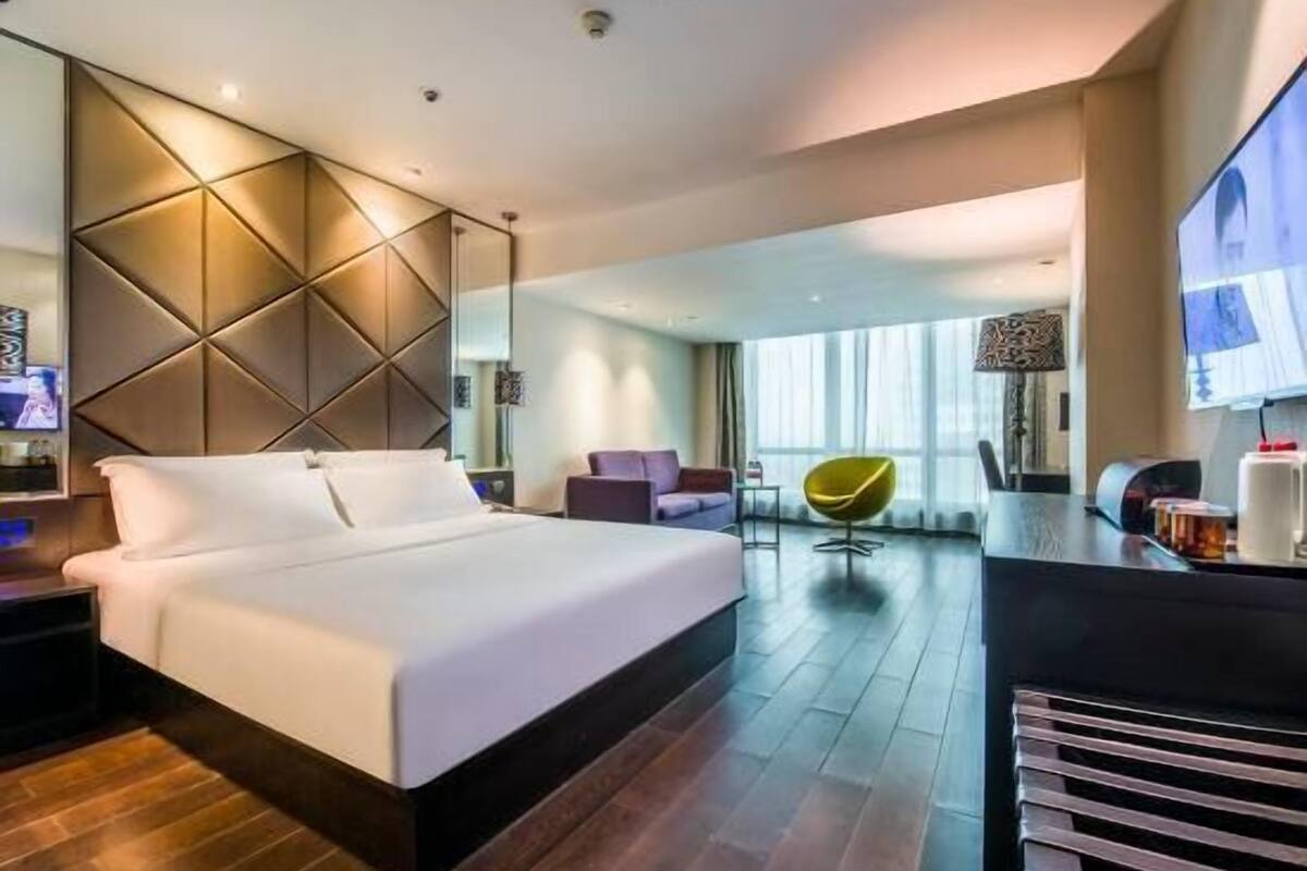 A modern hotel room with a large bed, a seating area with a chair, a TV, and a large window.