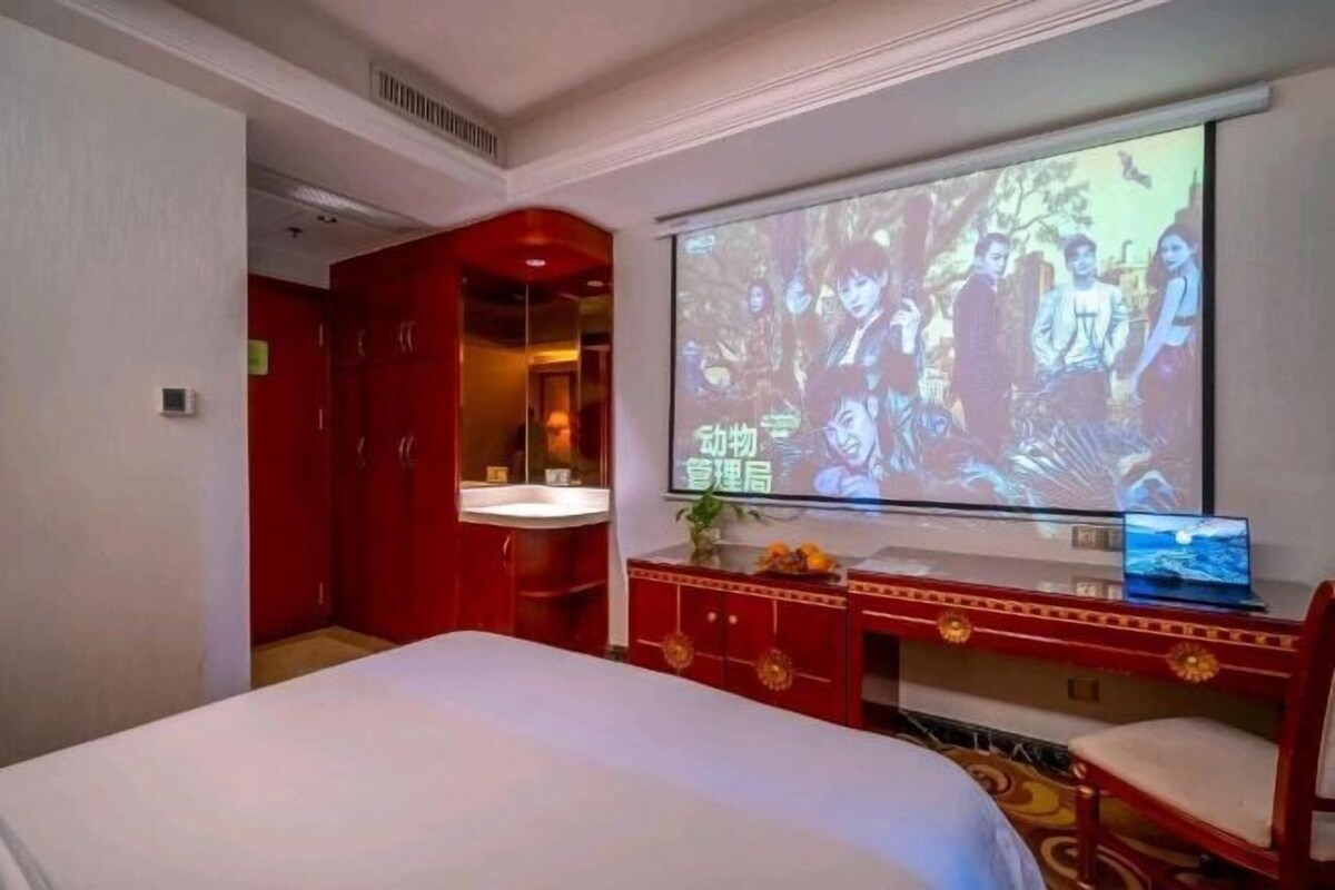 A hotel room with a bed, a TV screen displaying a movie, a desk with a laptop, and a red wardrobe.