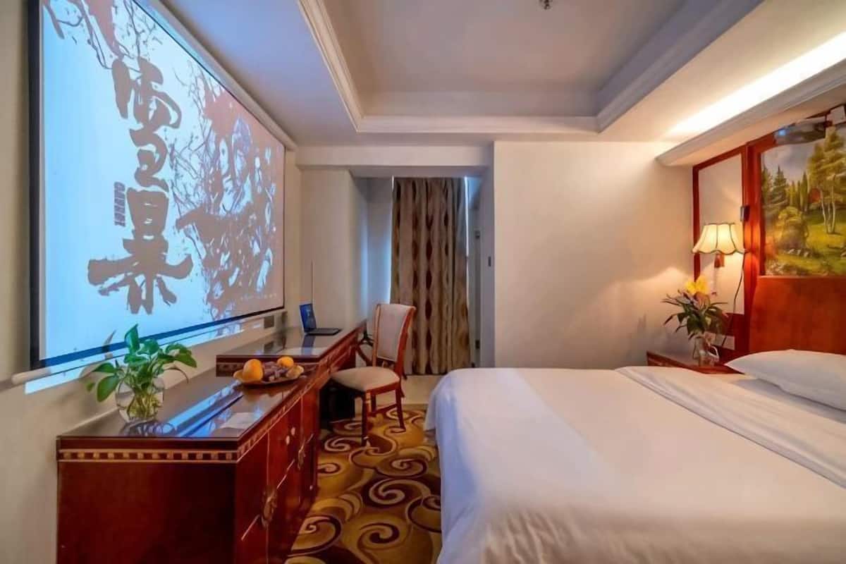 A hotel room with a bed, desk, chair, and a large screen displaying Chinese characters.