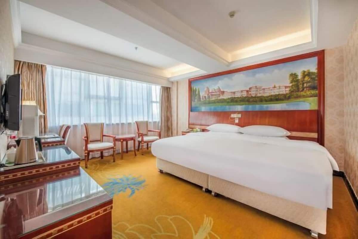 A hotel room with a large bed, a desk with a lamp, two chairs, a painting on the wall, and a window with curtains.