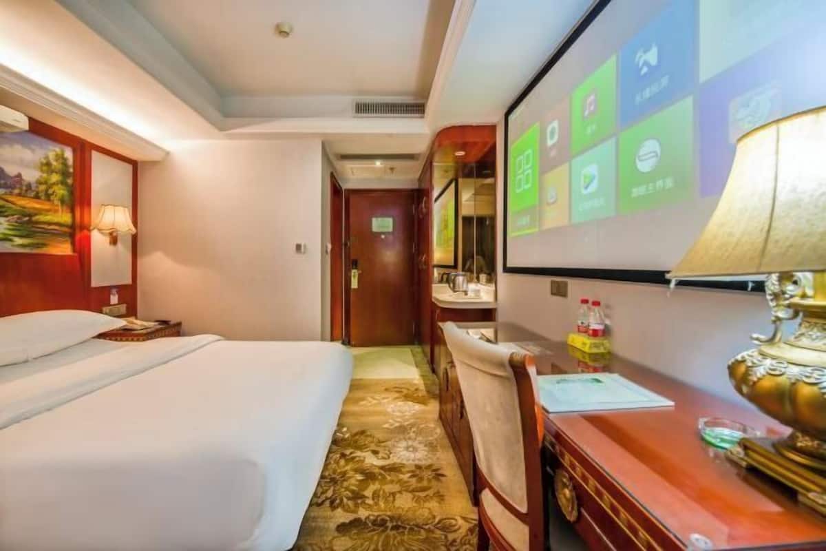 A hotel room with a bed, a desk, a chair, a lamp, a mirror, and a screen displaying room information.