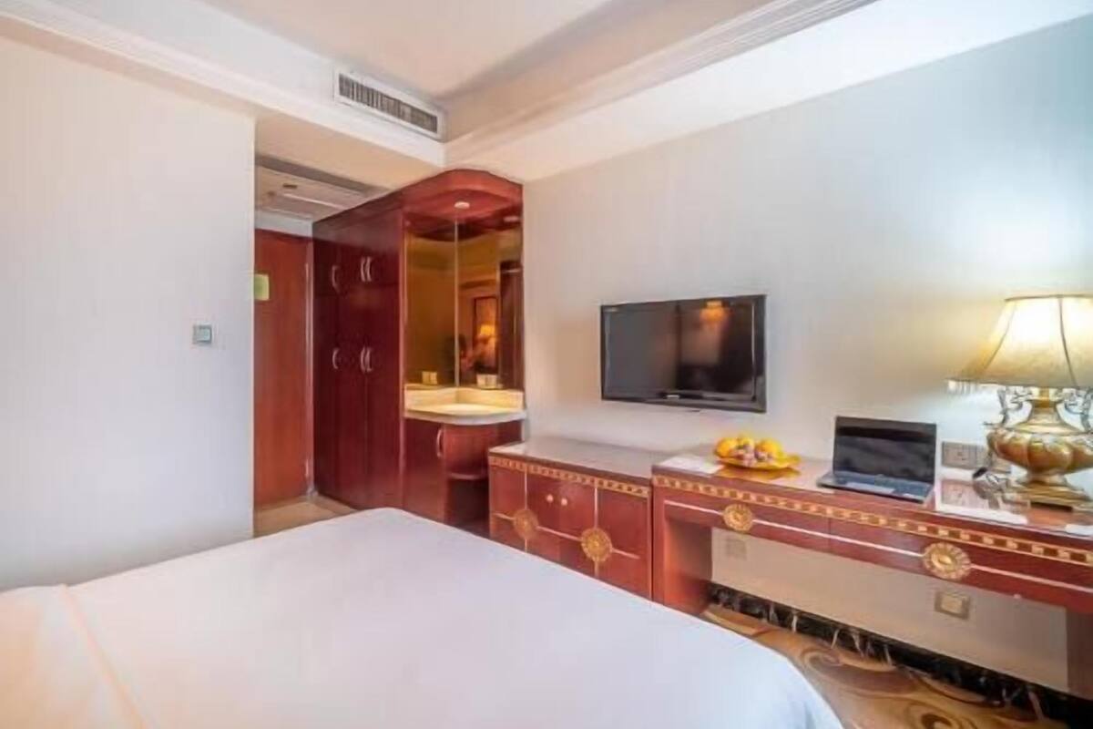 A hotel room with a bed, a television mounted on the wall, a desk with a lamp, and a wardrobe.