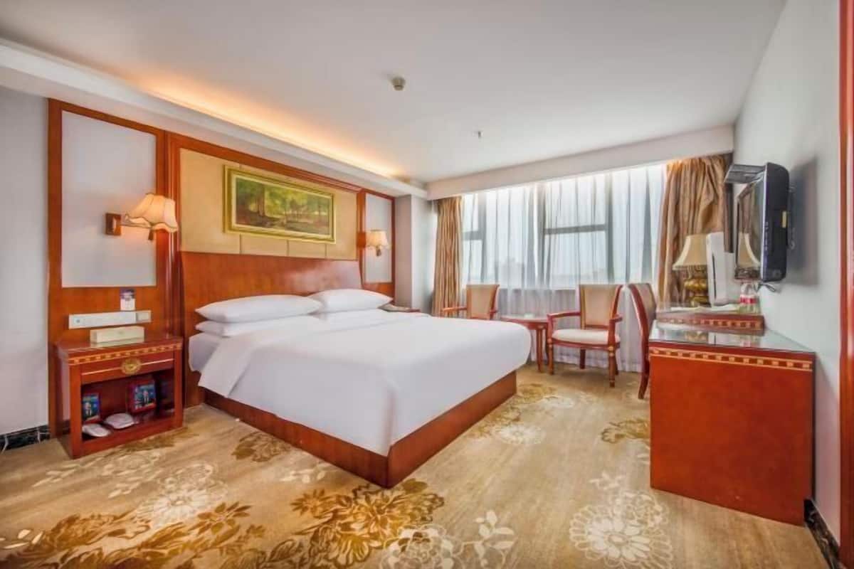 A hotel room with a large bed, a desk with a lamp, a television, and a window with curtains.