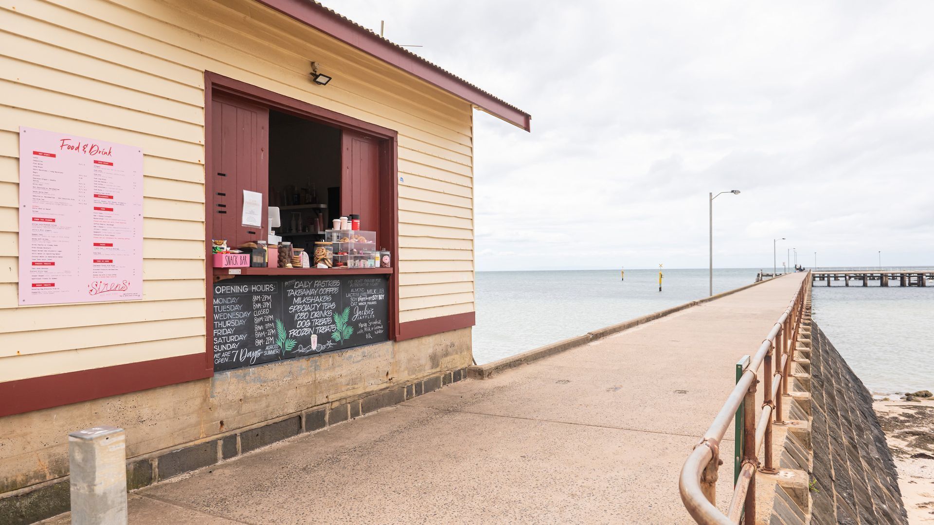 A seaside snack bar with a menu board, located next to a pier extending into the water.