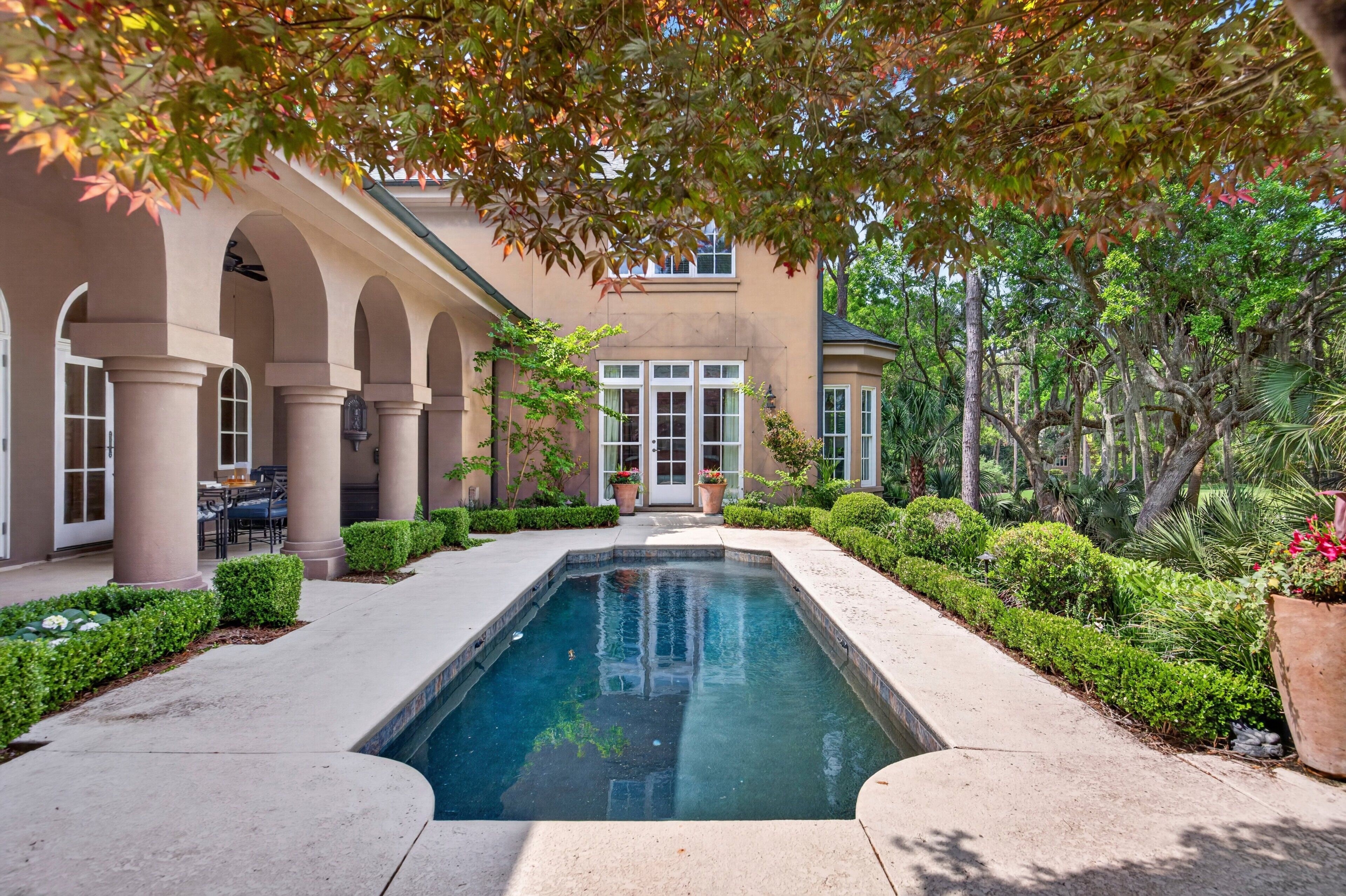 A pool with a surrounding patio, a building with arches, and lush greenery.