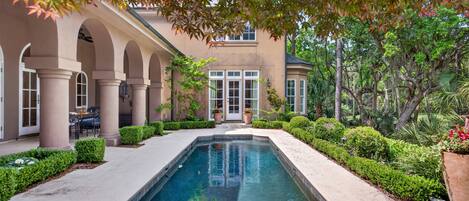 A pool with a surrounding patio, a building with arches, and lush greenery.