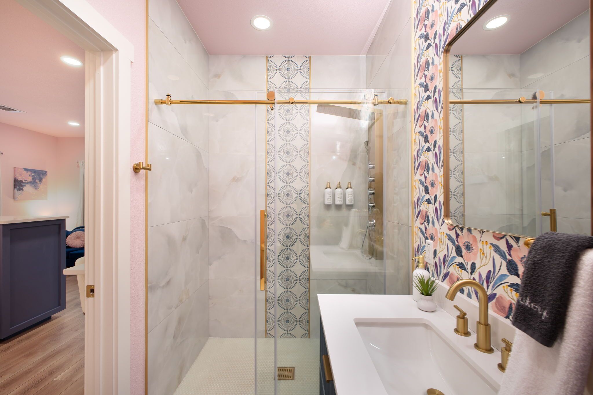 A modern bathroom with a glass shower enclosure, a white sink with gold fixtures, and a mirror with a floral backsplash.