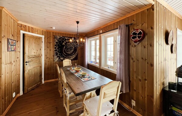 A wooden-paneled room with a dining table, chairs, a chandelier, and windows with curtains.
