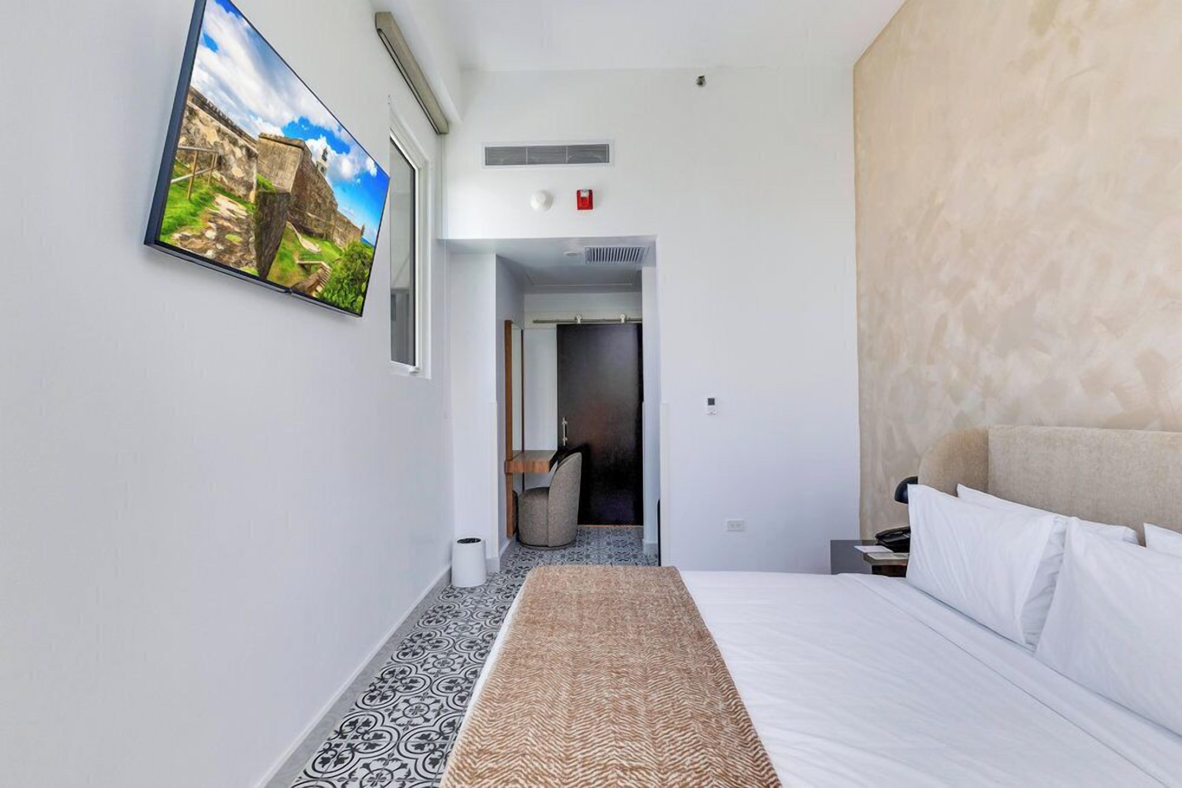 Quarto de hotel com cama, televisão fixada na parede, porta para um banheiro e piso estampado.