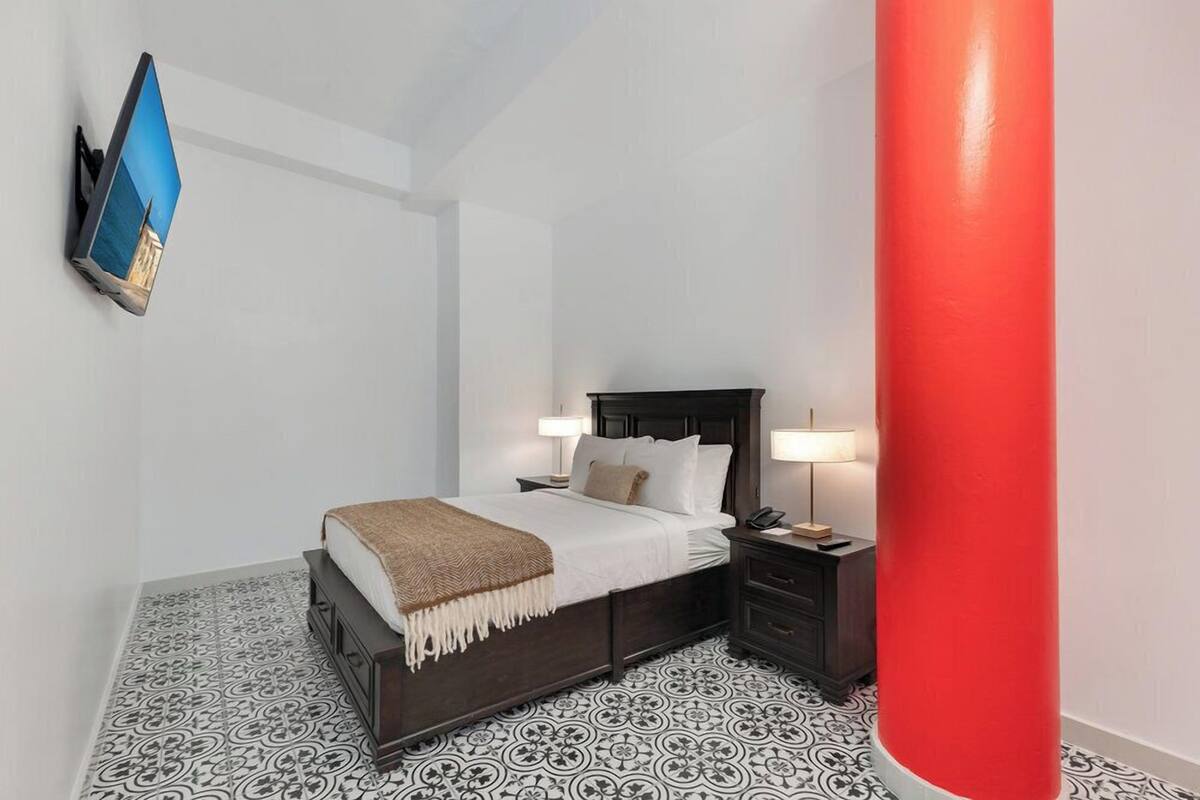 A bedroom with a bed, bedside tables, a TV mounted on the wall, and a red pillar.