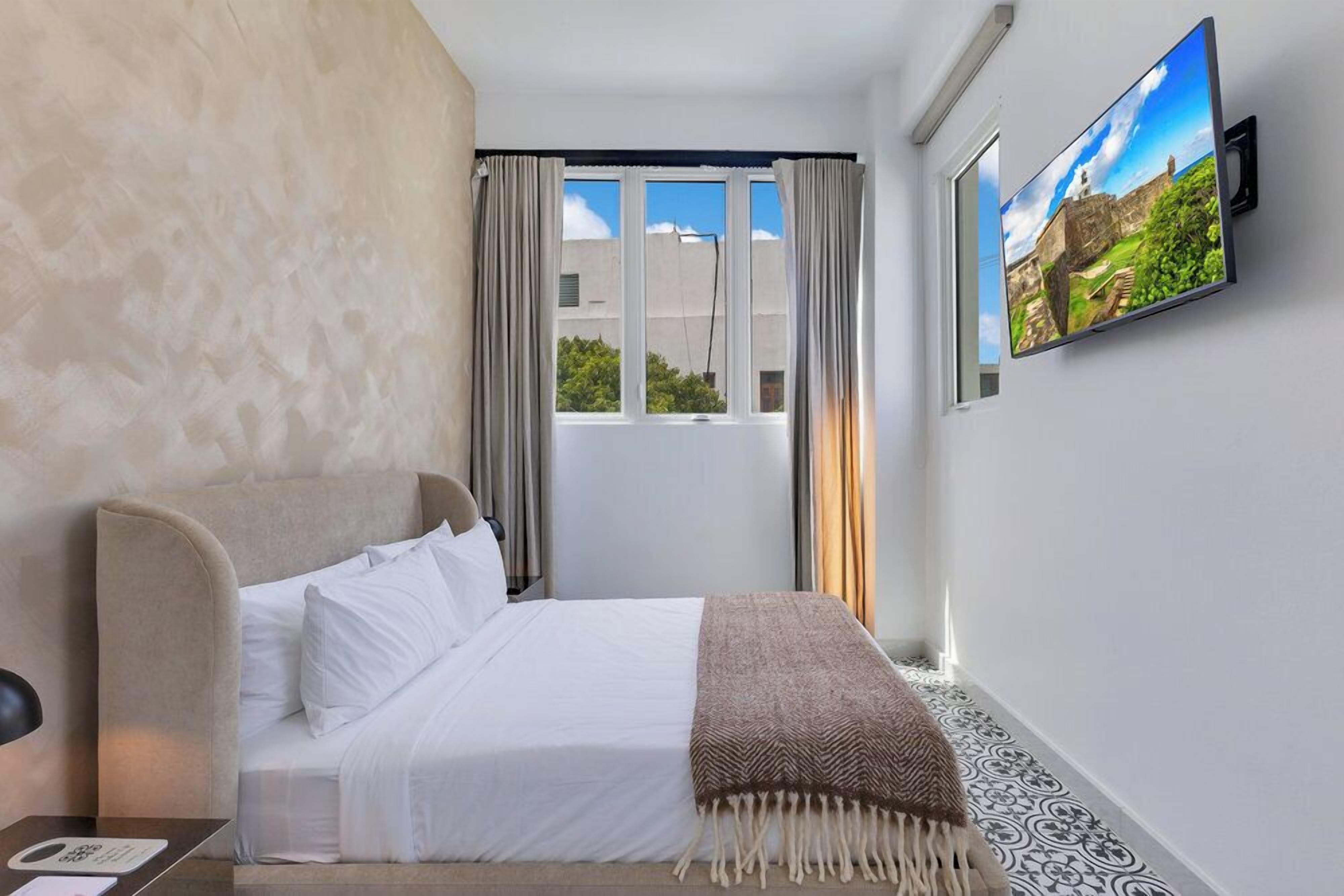Una habitación de hotel con una cama, televisión montada en la pared, ventana con cortinas y una alfombra estampada.