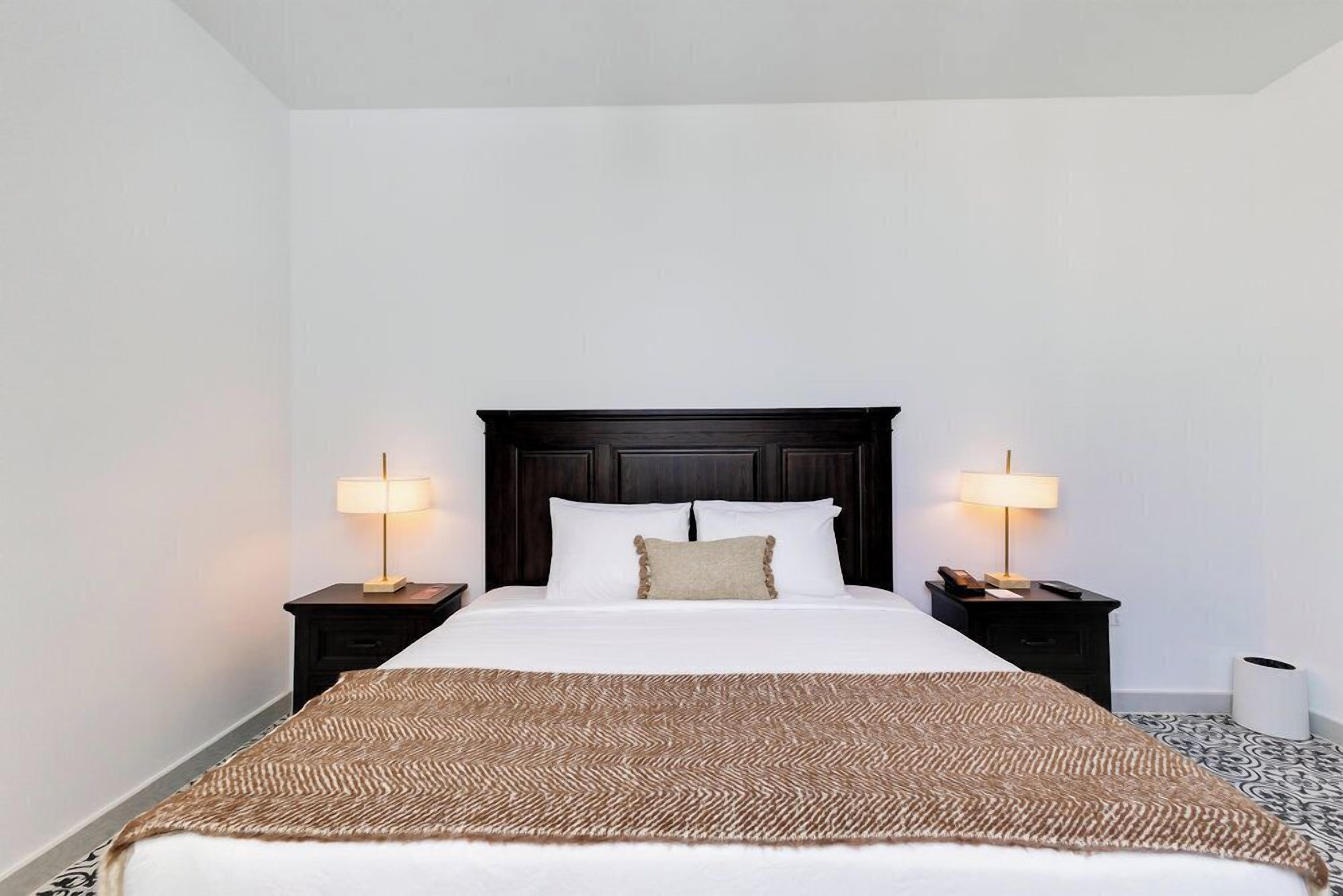Habitación de hotel con una cama grande, dos mesitas de noche con lámparas, un cabecero de madera oscura y una alfombra estampada.