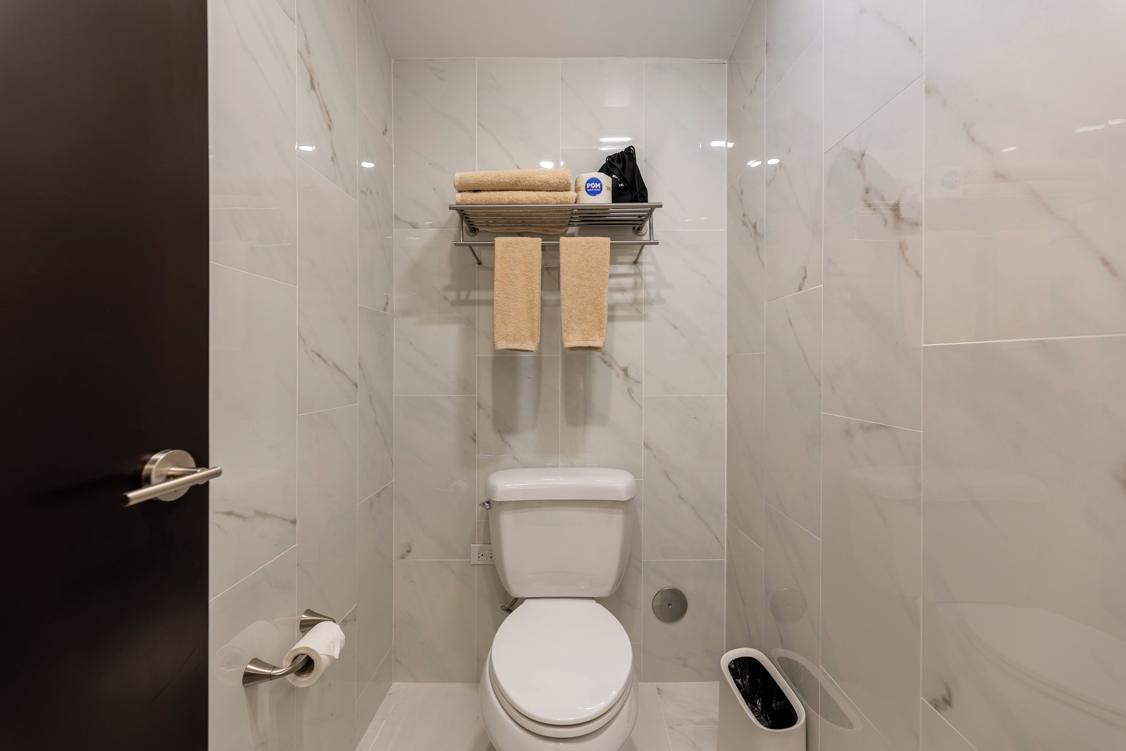 Un baño moderno con inodoro, estante con toallas, rollo de papel higiénico y cubo de basura.