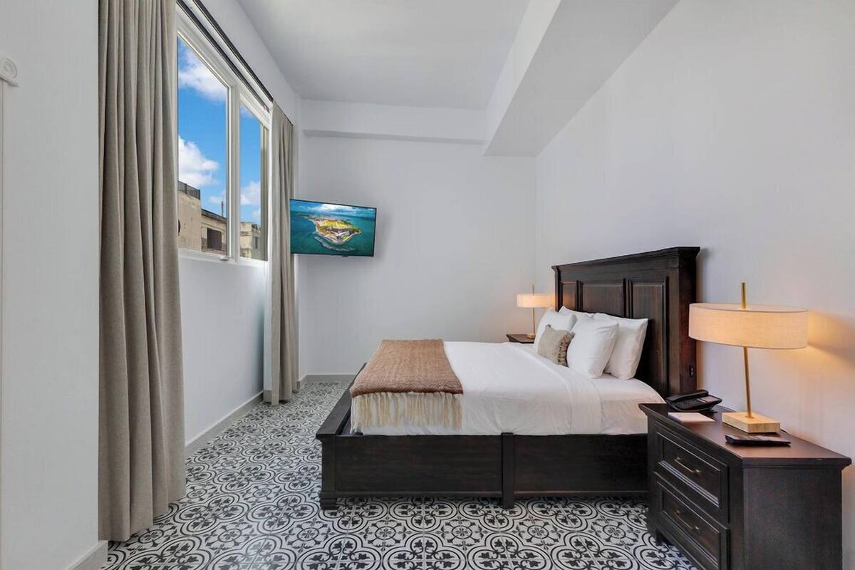 A hotel room with a bed, bedside lamps, a TV on the wall, and patterned floor tiles.
