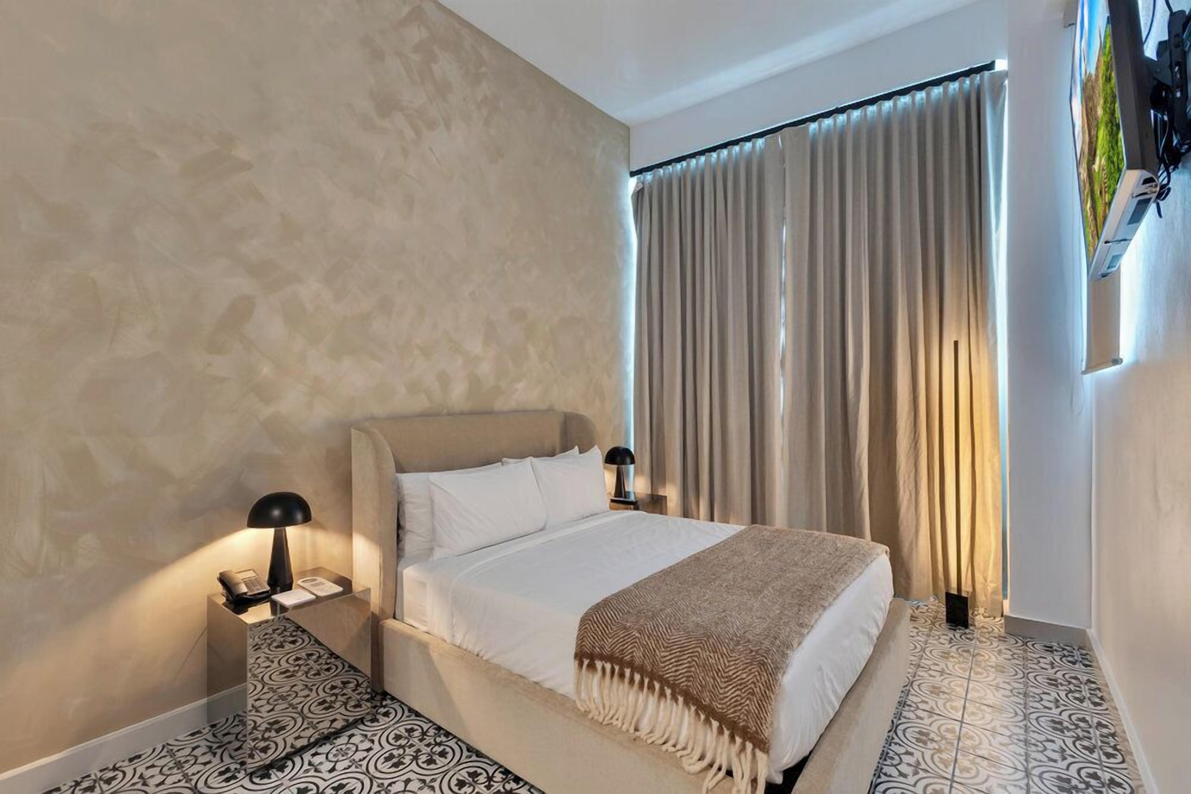 A hotel room with a bed, bedside lamps, a telephone, a window with curtains, and patterned floor tiles.