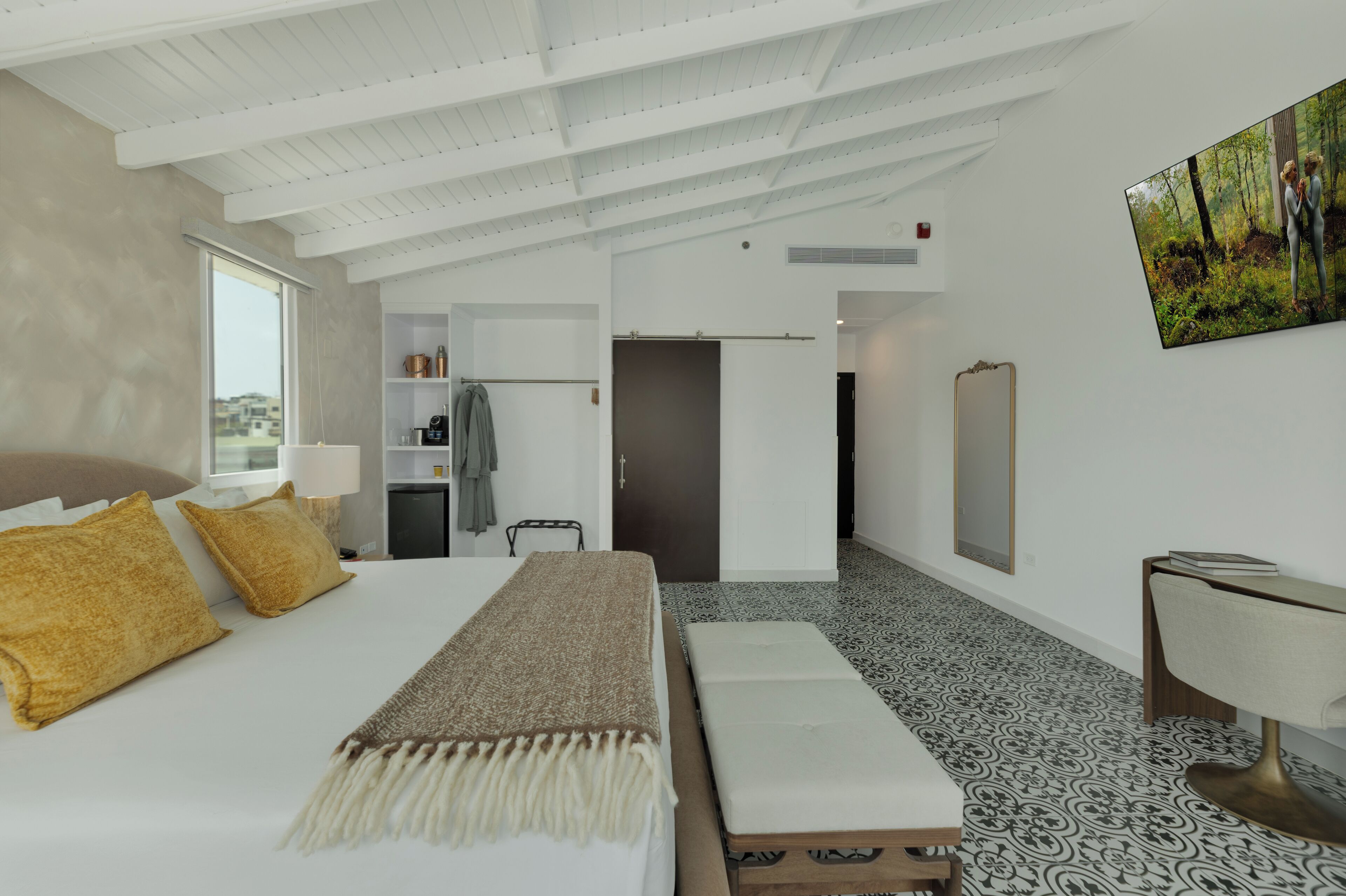 A modern hotel room with a large bed, a bench, a desk, and a wardrobe.