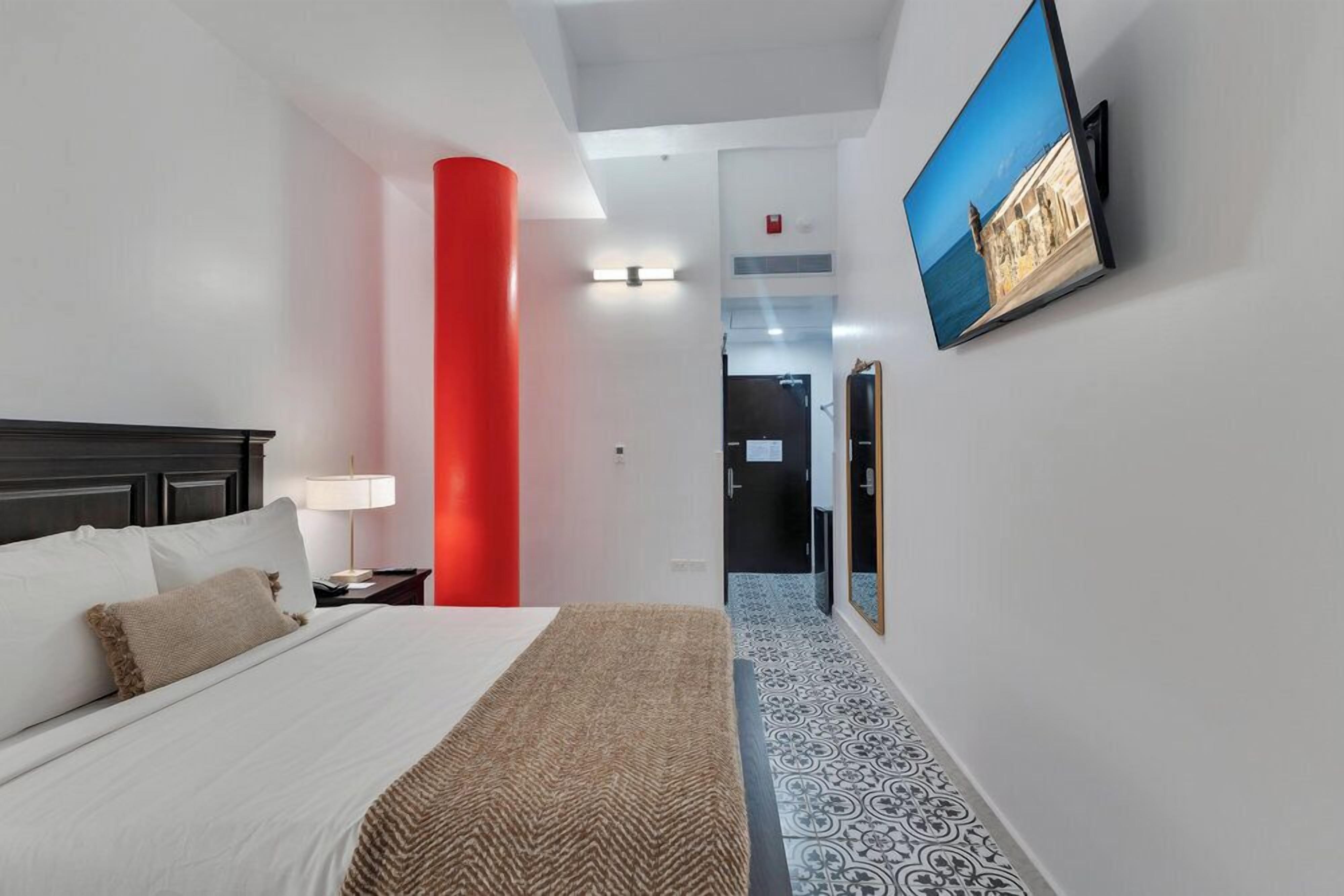 Habitación de hotel con cama, mesita de noche, lámpara, un pilar rojo, televisor montado en la pared y suelo estampado.