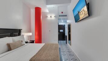 Habitación de hotel con cama, mesita de noche, lámpara, un pilar rojo, televisor montado en la pared y suelo estampado.